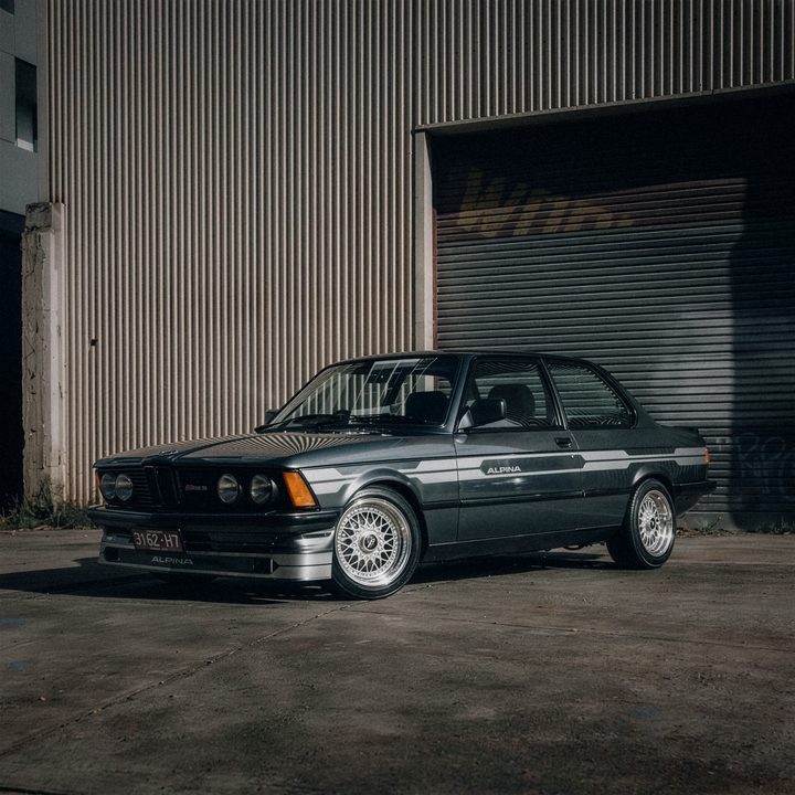 Dark grey BMW E21 with Alpina side stripes and silver mesh wheels, outside a warehouse roller door