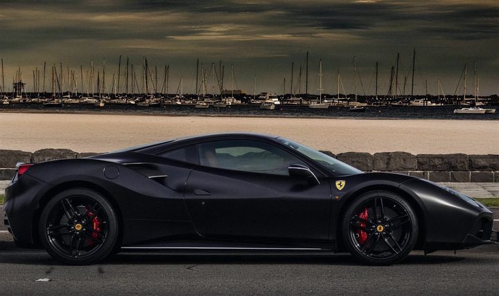 Matte black Ferrari 488 GTB on a seaside road with yachts behind