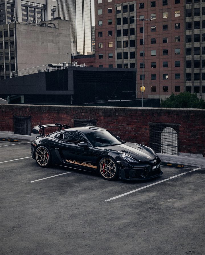 Black Porsche 718 Cayman GT4 RS with a gold side script, on a rooftop car park in the city